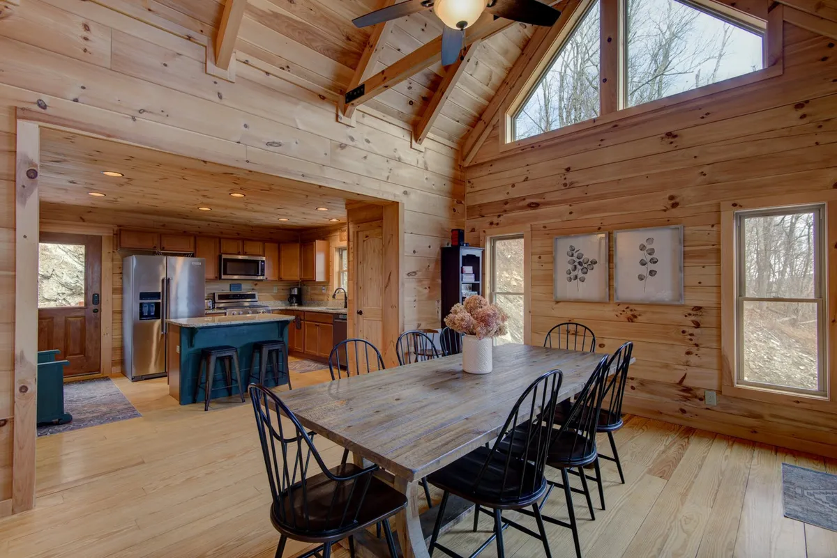 A spacious dining area with a large wooden table and chairs, adjacent to a modern kitchen with stainless steel appliances. Sunlight pours in through expansive windows, wooden ceiling beams add rustic charm, and the kitchen area is clearly stocked and functional.