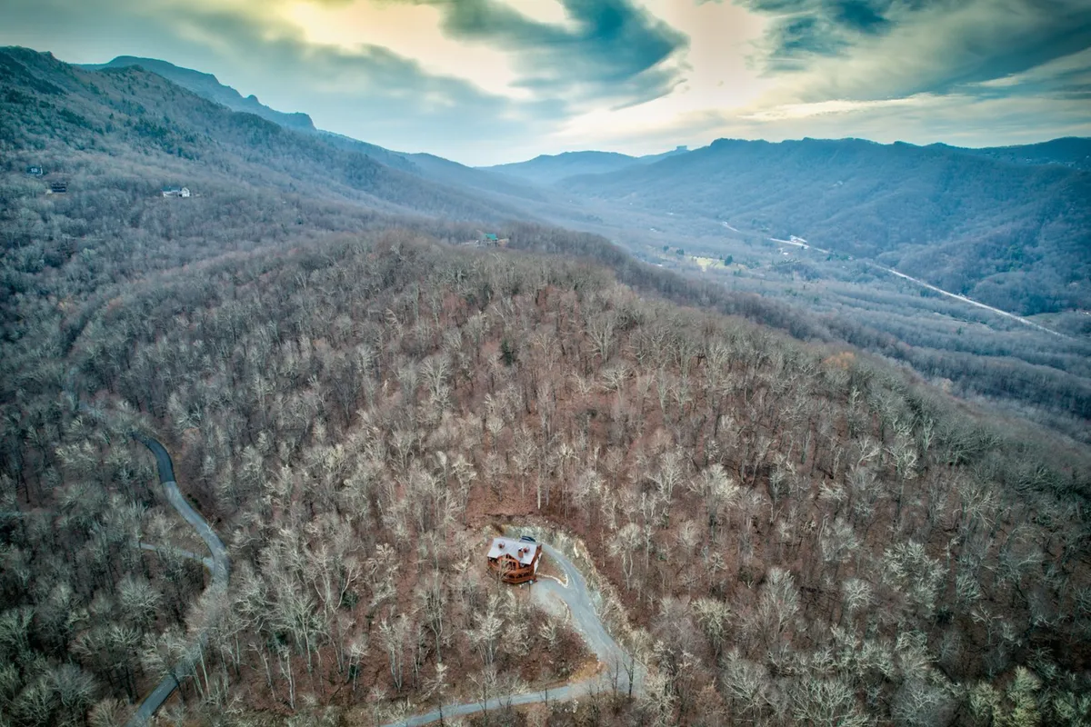 The home is embraced by a dense forest, with a dramatic mountain range visible in the background. The composition emphasizes the secluded atmosphere while showcasing the immediate access to nature and adventure right beyond the doorstep.