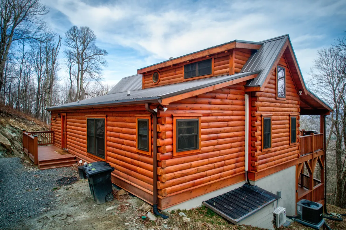 A view of the log cabin’s exterior with an inviting deck surrounded by lush trees, showcasing the seamless connection between indoor comfort and the beauty of nature. The spacious deck suggests areas for outdoor gatherings, relaxation, and enjoying the scenery.