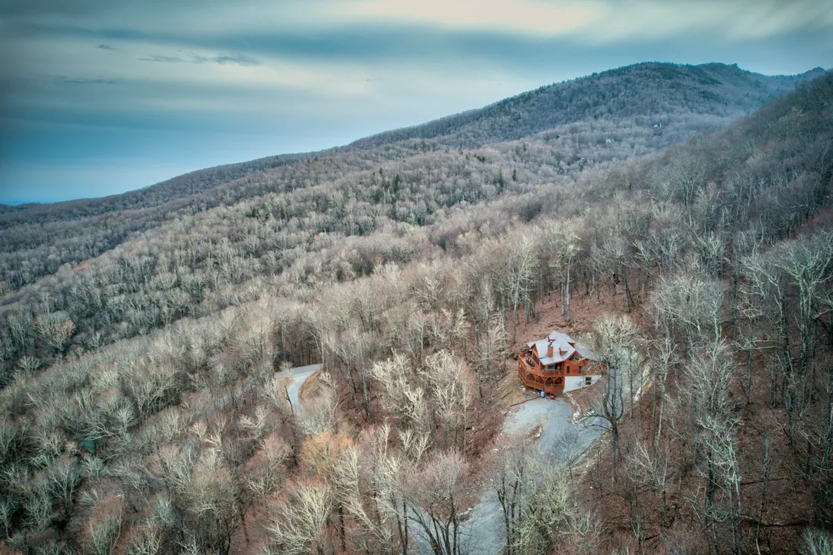 A broad view of the house, surrounded by mature trees and a winding driveway that hints at seclusion within nature. The expansive woodland landscape evokes the sense of plentiful trails and outdoor exploration opportunities surrounding the property.