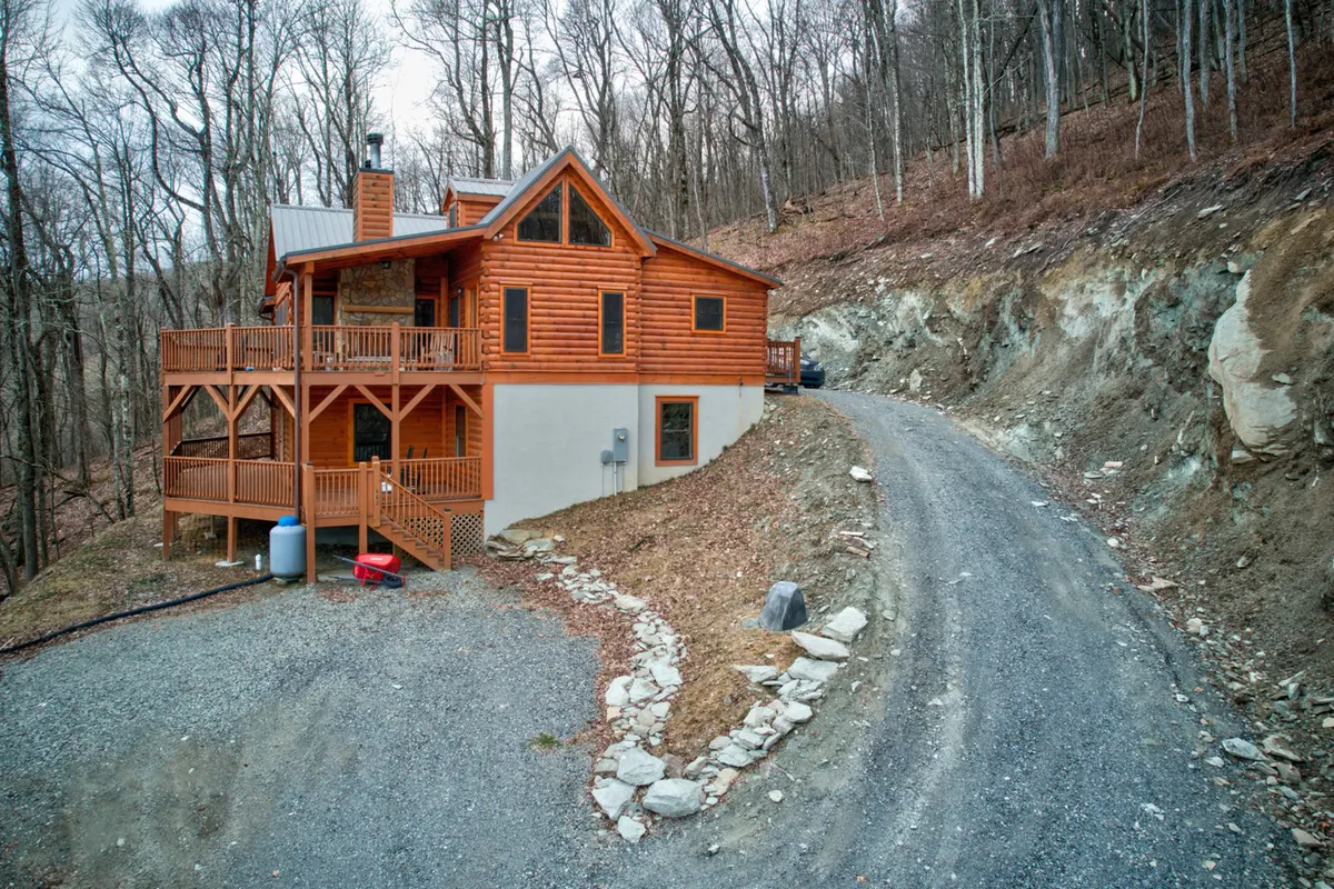 This image features the exterior of a cozy wooden cabin surrounded by tall trees, with a gravel driveway and a spacious deck area. The scene evokes a sense of seclusion and immersion in nature, ideal for outdoor activities and relaxation. The setting is perfect for exploring trails with pets and offers a welcoming entry for guests and their furry companions.
