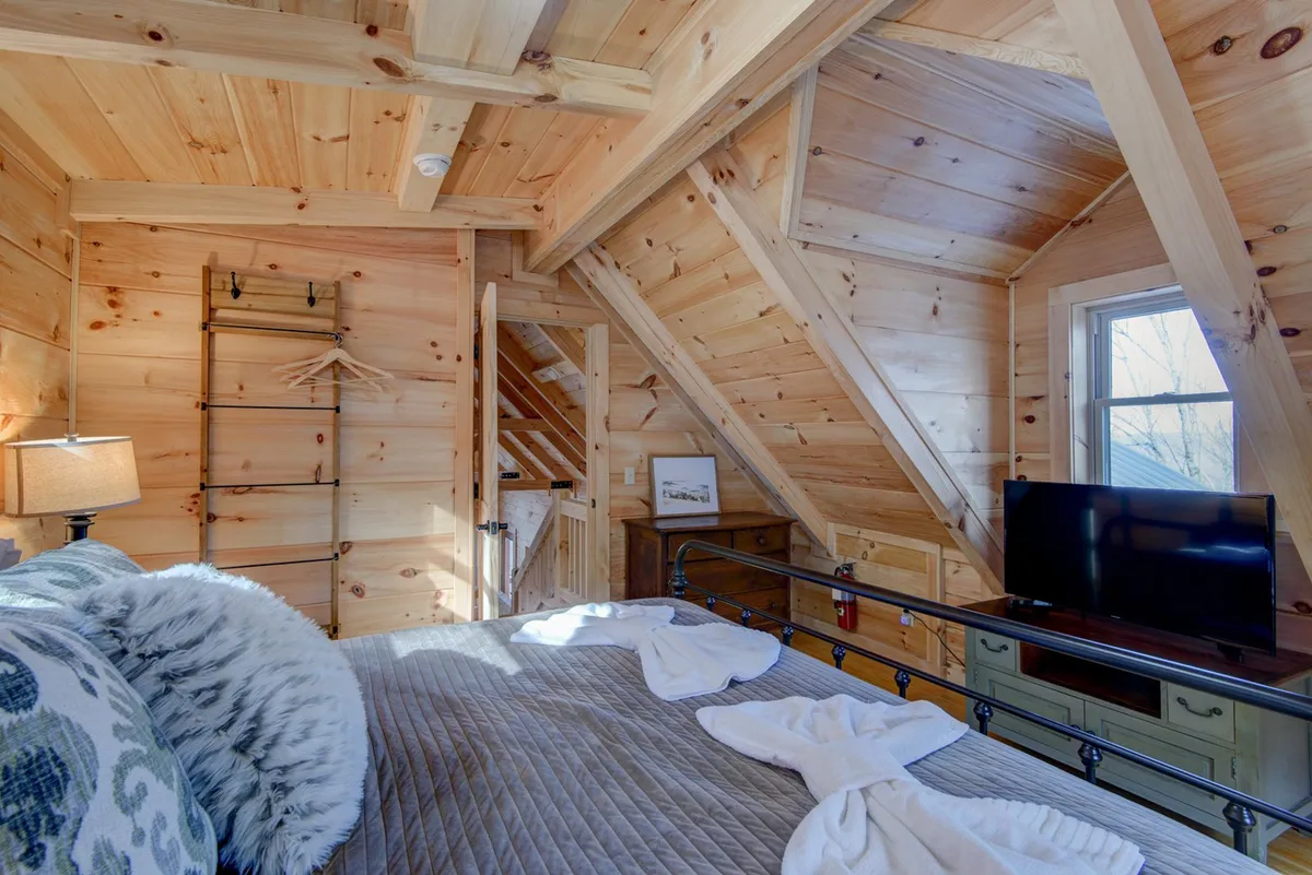 An upper-level bedroom featuring exposed wooden beams, a bed adorned with decorative pillows, a small lamp, a TV, and a window providing natural light. The warm tones and cozy furnishings create an inviting atmosphere ideal for a private retreat.