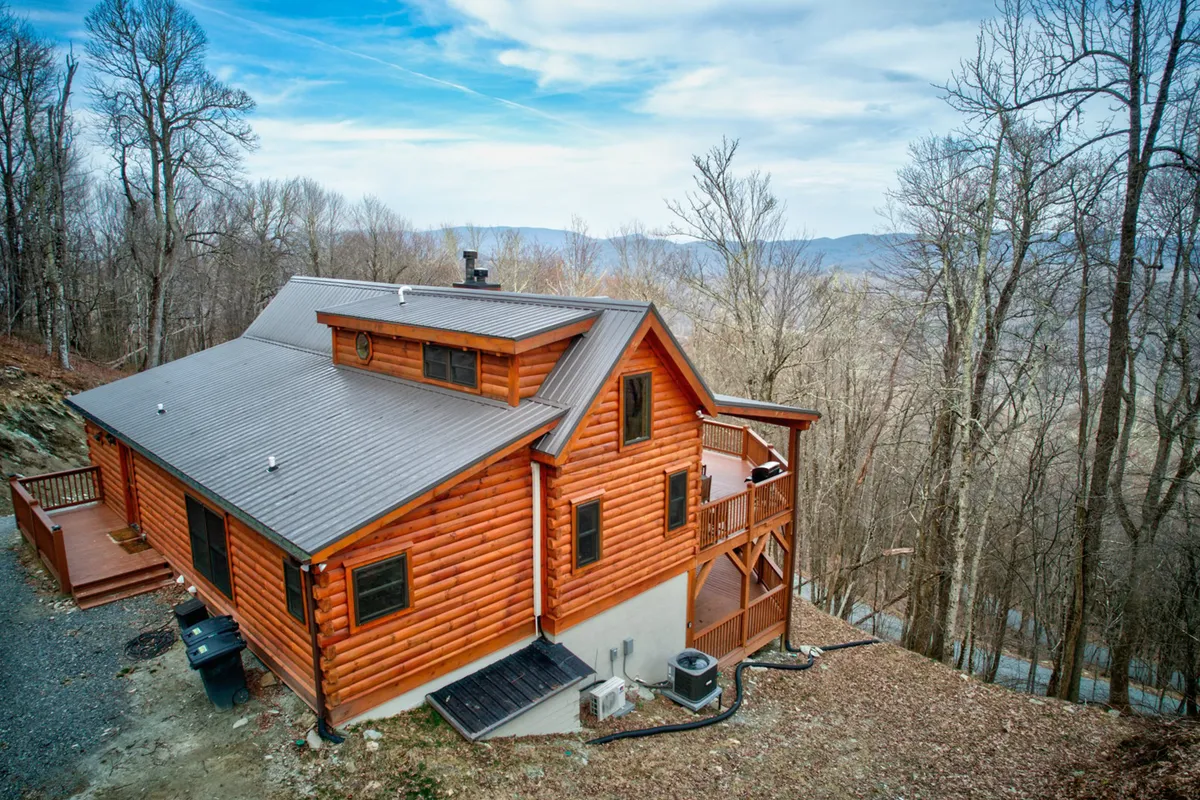 This exterior shot features the log cabin structure with a visible deck area, surrounded by trees and offering a clear glimpse of distant mountain views. The image is bathed in inviting, natural light, suggesting peaceful mornings and memorable evenings on the deck.