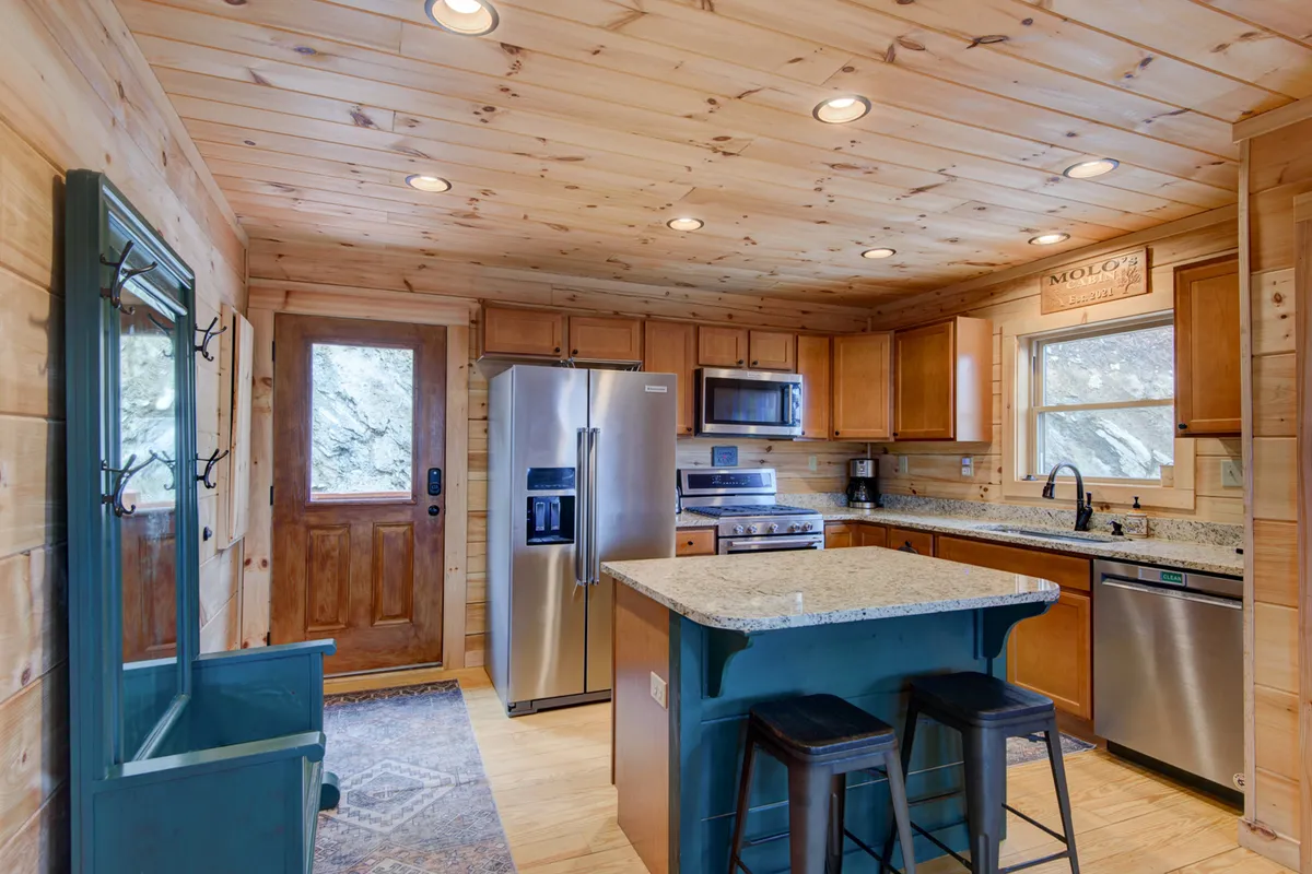 A bright, functional kitchen with a central island, stainless steel appliances, rich wooden cabinetry, and a natural wood ceiling. The space feels ready for group meal prep, with ample counter space and utensils on display.