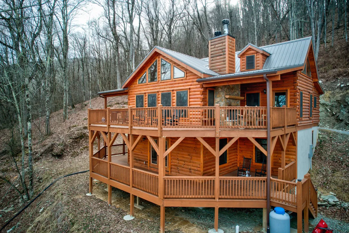 The image showcases a charming log cabin surrounded by lush trees, featuring a spacious covered deck with inviting seating. The sunset casts a warm glow over the wood exterior, creating a cozy and welcoming atmosphere. The deck overlooks tranquil mountain scenery, hinting at peaceful mornings and relaxing evenings immersed in nature.