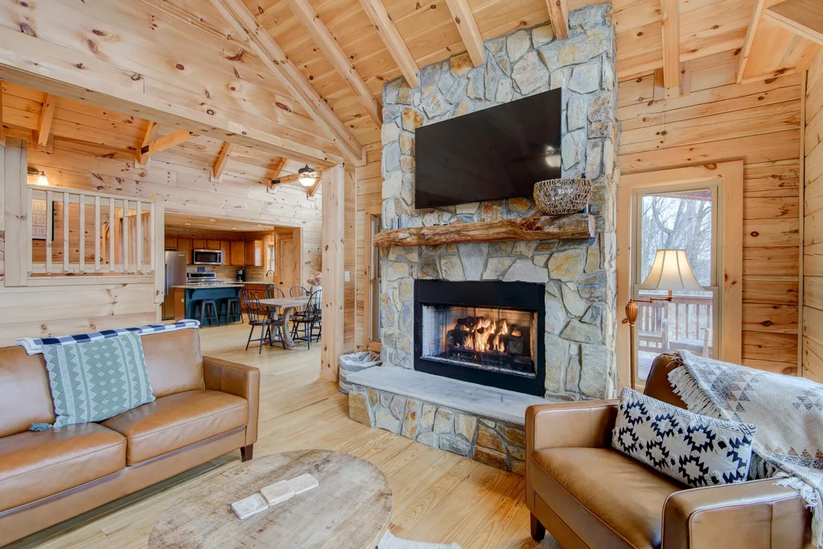 A warm, open-concept living area featuring a striking stone fireplace, a plush leather sofa, and an inviting wooden ceiling. Large windows bathe the space in natural light, with the dining area visible in the background, suggesting a seamless flow between living and dining spaces.