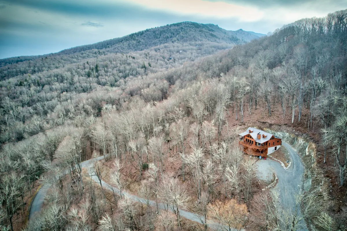 A rustic house is nestled at the end of a curved driveway, surrounded by towering mountains and lush woodland. The peaceful setting hints at abundant outdoor recreation, including nearby hiking trails and nature walks. The expansive views suggest a gateway into the surrounding wilderness.