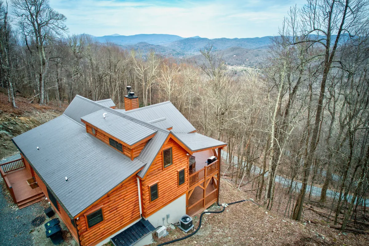 A log cabin structure nestled within a tranquil mountain setting, surrounded by dense forest and distant mountain views. The image captures both the intimacy of the home’s immediate natural environment and the grandeur of the mountainous landscape beyond, suggesting direct access to hiking and exploration right outside the door.