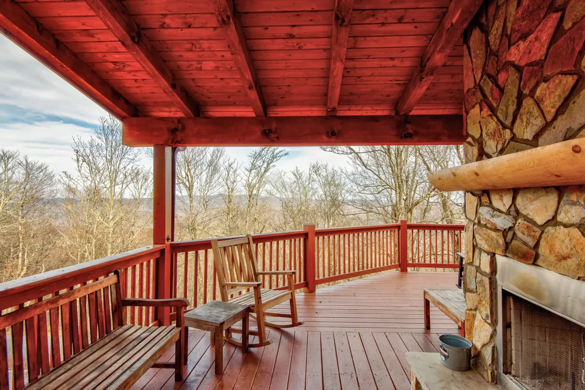 This image shows a serene covered deck with wooden benches, a substantial stone fireplace, and a wooden railing looking out over surrounding trees. The ambiance is tranquil, offering a prime spot to enjoy morning coffee, socialize with friends, or watch the sunset.
