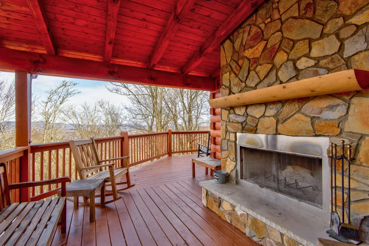This image captures a warm and inviting covered wooden deck with a prominent stone fireplace, red ceiling accents, and classic rocking chairs arranged for relaxation. The scene exudes coziness and suggests a space ideal for gathering, enjoying fireside chats, and appreciating the fresh air.